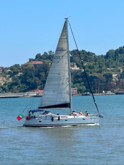 A sailboat with a partially unfurled sail on calm water, with a coastal town and hills in the background. 3