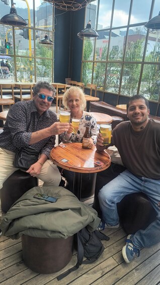 Three people sitting at a wooden table with drinks, in a cafe with large windows and outdoor seating. 3