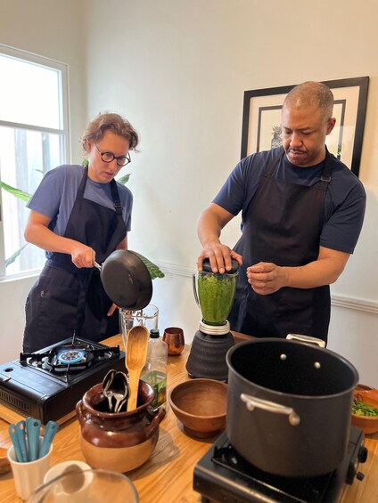 Two people preparing a green mixture in a kitchen with a stove, pots, and wooden utensils. 5