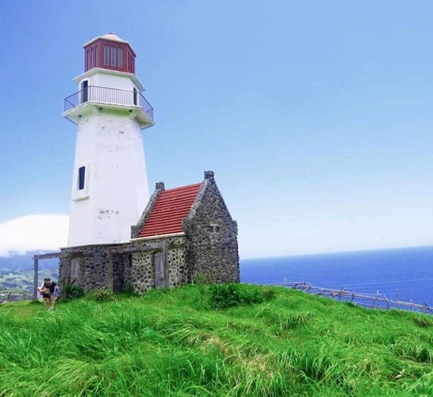 A lighthouse with a red roof and a white tower, adjacent to a stone building with a red roof, set against a clear blue sky and ocean. 1