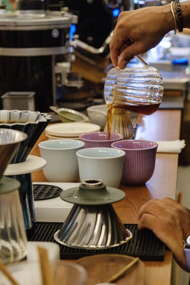 A person pouring a liquid from a glass jug into a metal strainer on a wooden table. 4