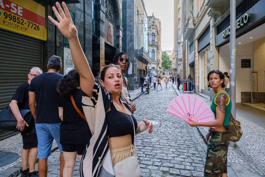 A cobblestone street with pedestrians, a woman holding a pink fan, and storefronts. 5