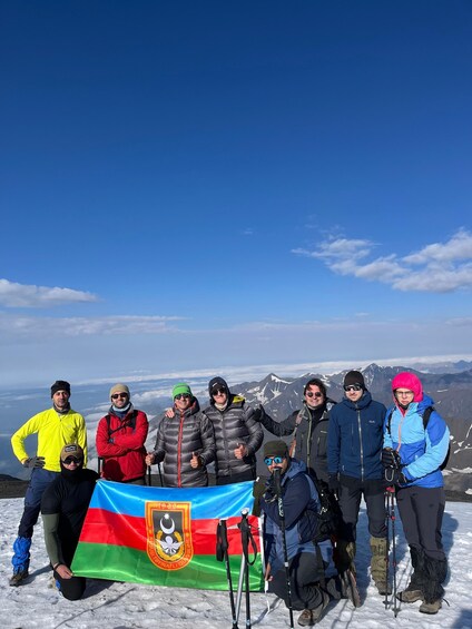 Un groupe de personnes se trouve sur une montagne enneigée, un drapeau est visible, et des montagnes se dessinent à l'arrière-plan, sous un ciel dégagé. 4