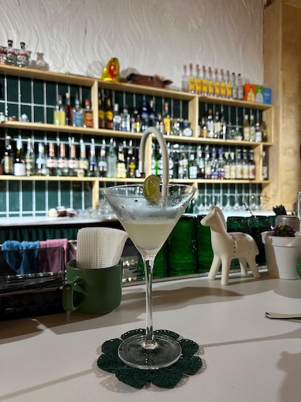 A martini glass with a lime wedge on a doily, placed on a bar counter with a green coaster. 4
