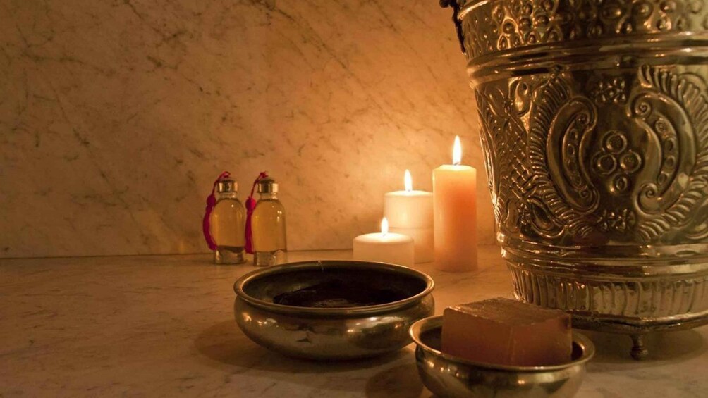 A brass oil lamp, two glass bottles with red tassels, a lit candle, and a metal bowl on a marble surface. 1