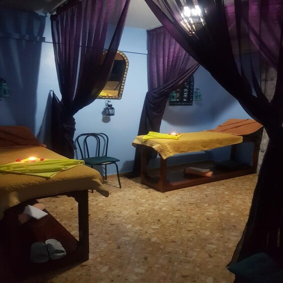 A room with two beds, a chair, a small table with a candle, and purple curtains. 2