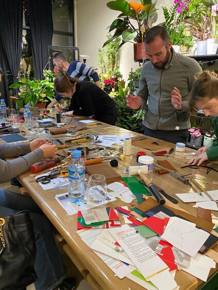 A group of people at a table with craft supplies, papers, and tools, surrounded by potted plants. 2