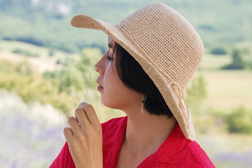 A person wearing a straw hat and a red polka dot blouse, standing outdoors with a blurred background of greenery and purple flowers. 2