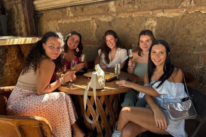 Een groep mensen zit buiten rond een houten tafel, met drankjes in hun handen. 1