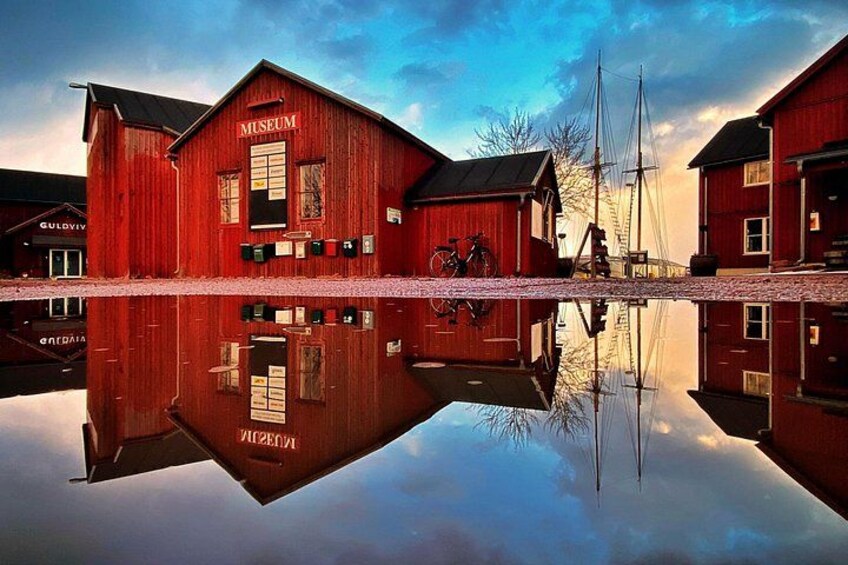 Een rood houten gebouw met de aanduiding 'Museum', met een fiets ervoor geparkeerd, weerspiegeld in een rustig waterlichaam. 2