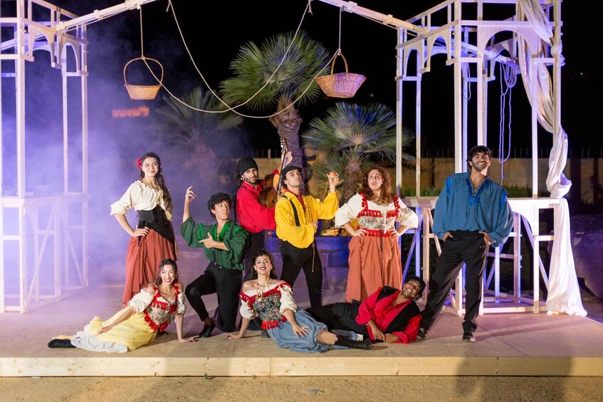 A group of people in traditional costumes on a stage with a white structure and palm trees in the background. 2