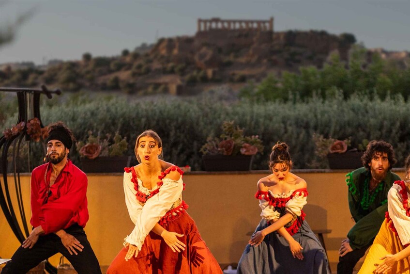 A group of people in period costumes on a stage with a backdrop of a hill and a building. 5
