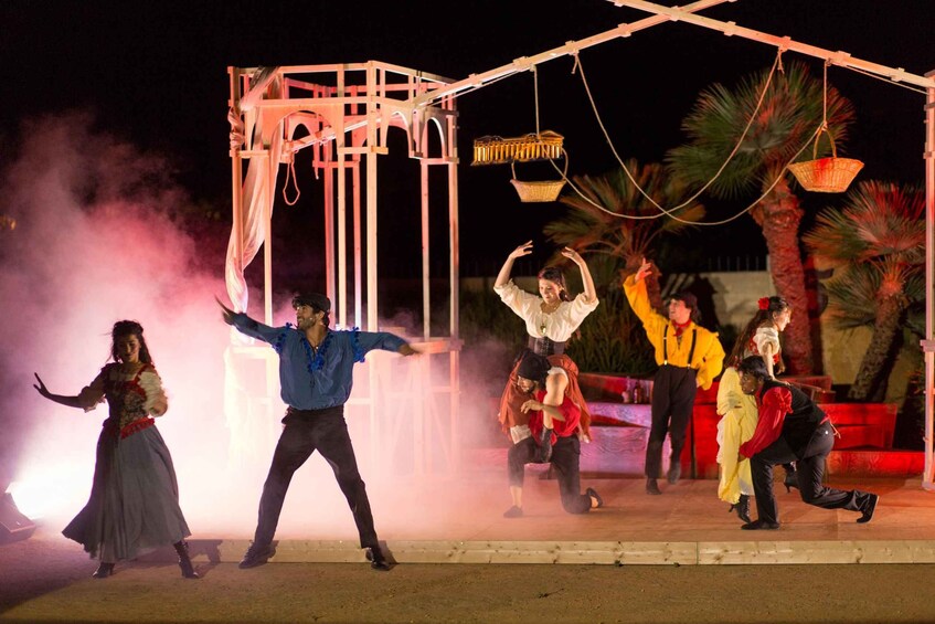 A nighttime outdoor performance with dancers in period costumes, a wooden structure, and palm trees. 1