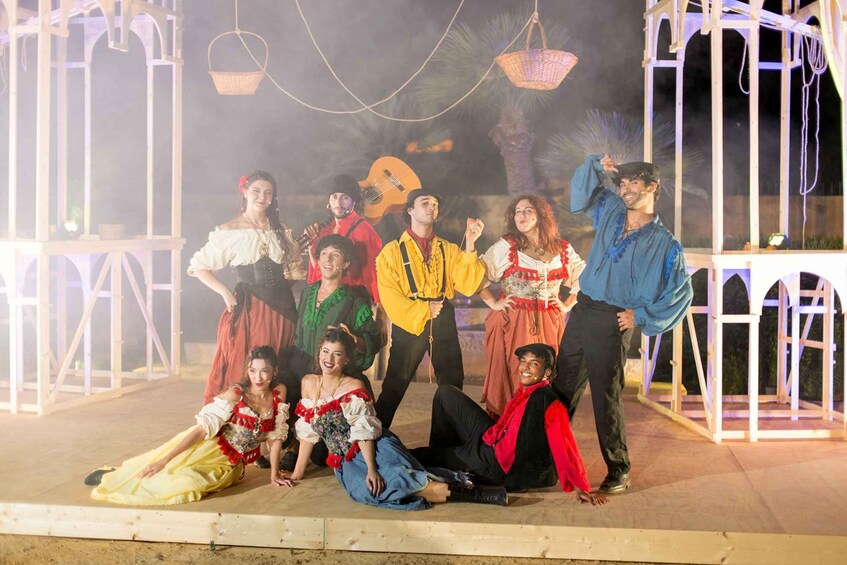 A group of people in traditional costumes on a stage with a white gazebo-like structure in the background. 3
