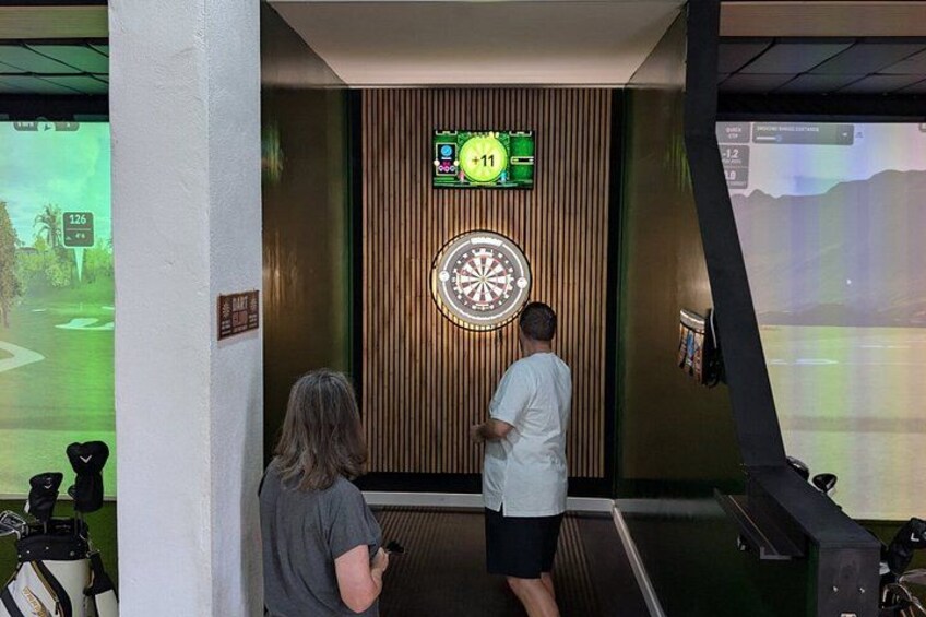 Two people playing darts at an indoor facility with a dartboard and a digital display. 1