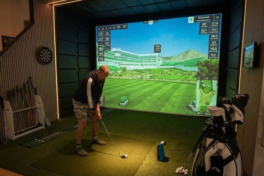 En person som spelar golf inomhus med hjälp av en virtuell golfbana som visas på en stor skärm. 1