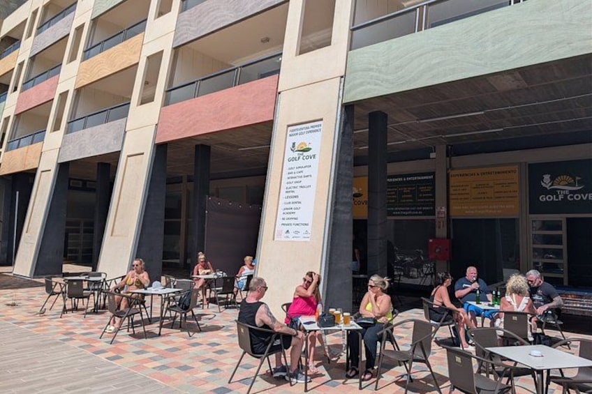 Un caffè all'aperto con tavoli e sedie, un edificio con un cartello e persone sedute che mangiano. 2