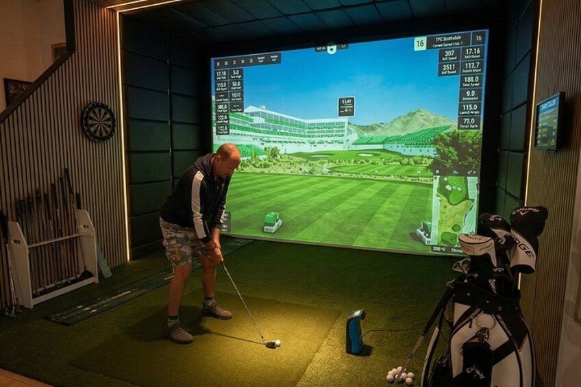 Una persona gioca a golf in un ambiente interno, con un percorso virtuale proiettato su uno schermo grande. 1