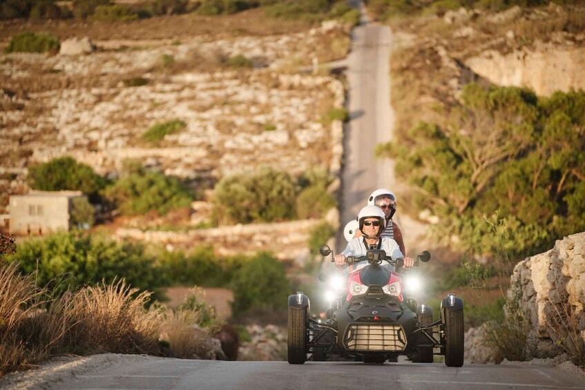 Two people on an ATV on a road with a rocky hillside in the background. 2