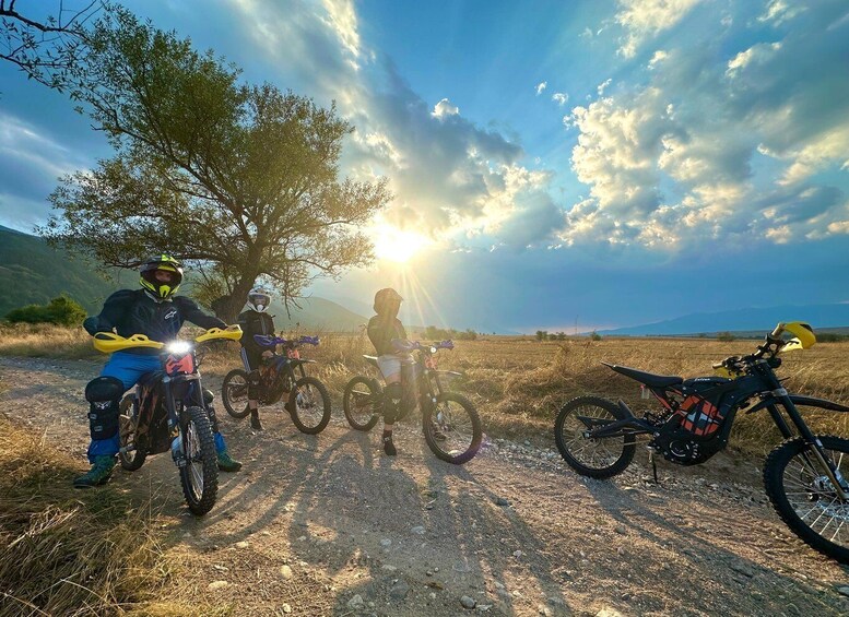 Three people on mountain bikes on a dirt path with a tree and a sunset in the background. 1