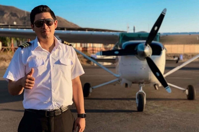 A person in a white uniform with a black belt and a watch, standing on a tarmac with a small propeller plane in the background. 2