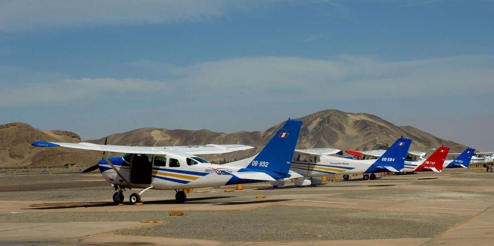 A row of small aircraft parked on a tarmac with mountains in the background. 5