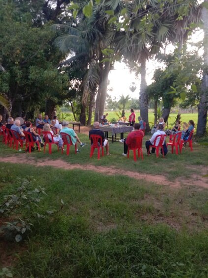 A group of people seated on red plastic chairs outdoors, gathered around a table with a tablecloth. 2