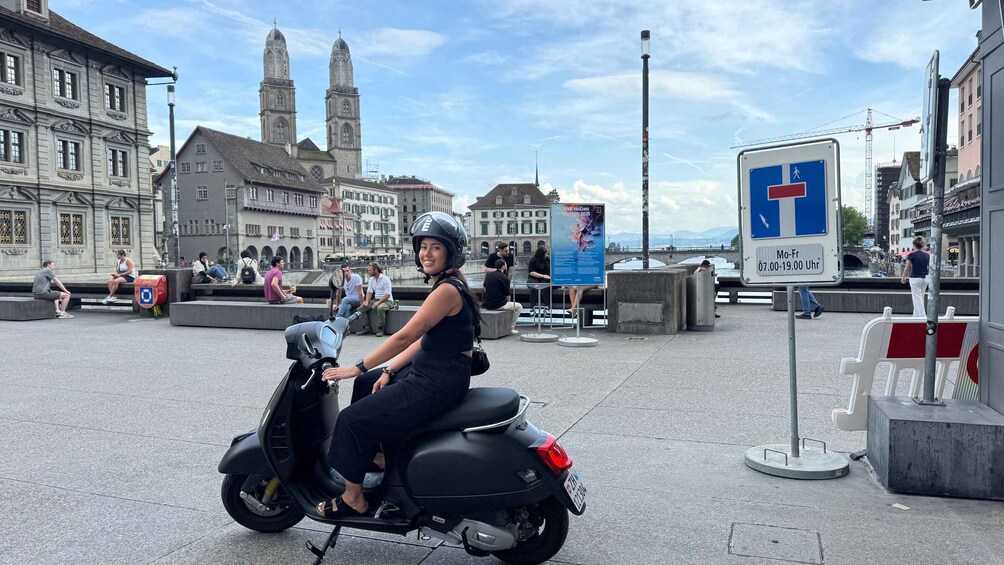 A person on a scooter in a public square with historic buildings and a river in the background. 3