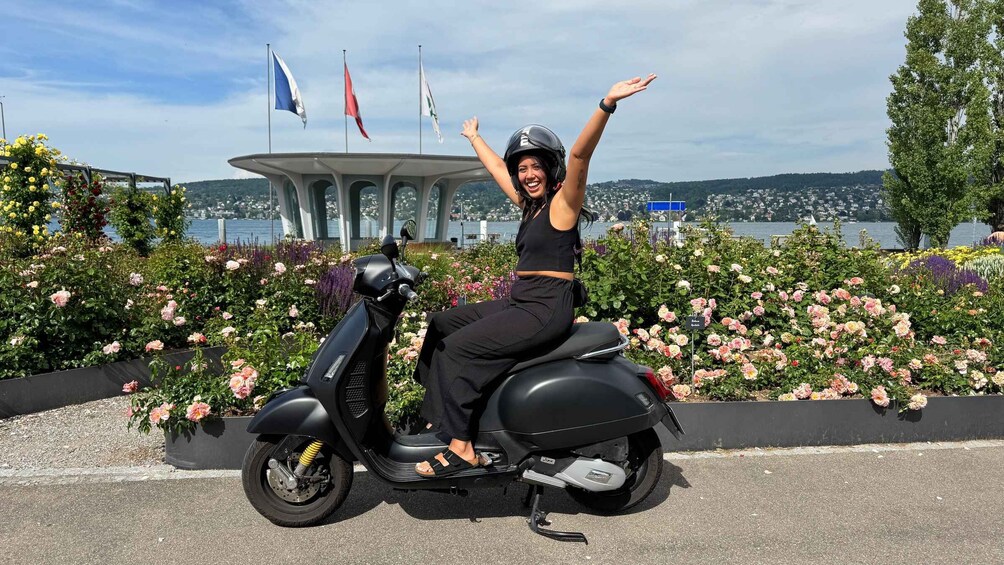 A person on a scooter with raised arms, surrounded by flowers and flags, with a body of water and buildings in the background. 1