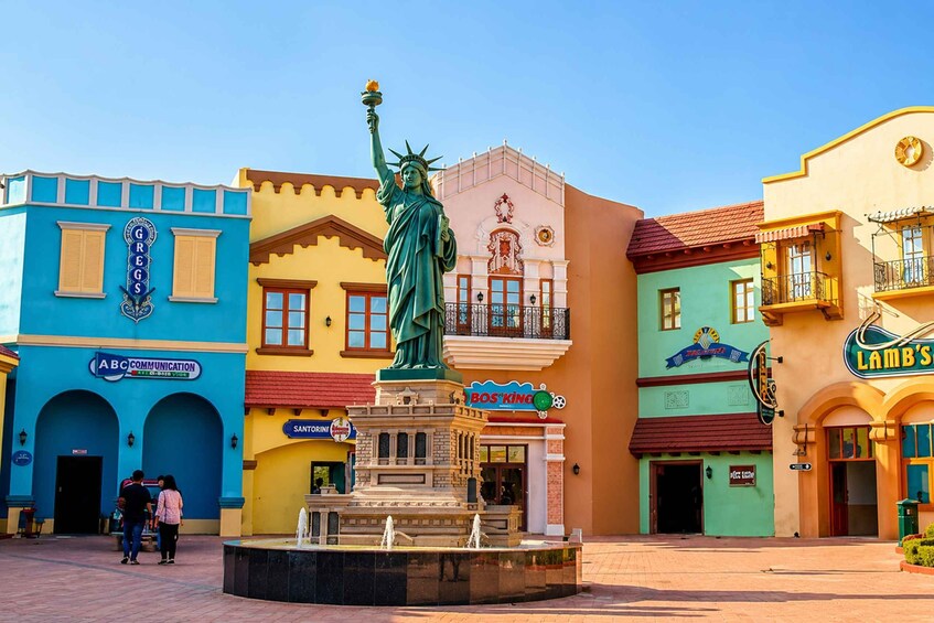 A colorful street with a statue of the Statue of Liberty, a fountain, and various shops. 1