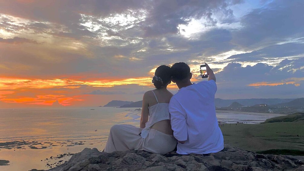 A couple sitting on a rocky outcrop overlooking a coastal landscape during sunset. 1