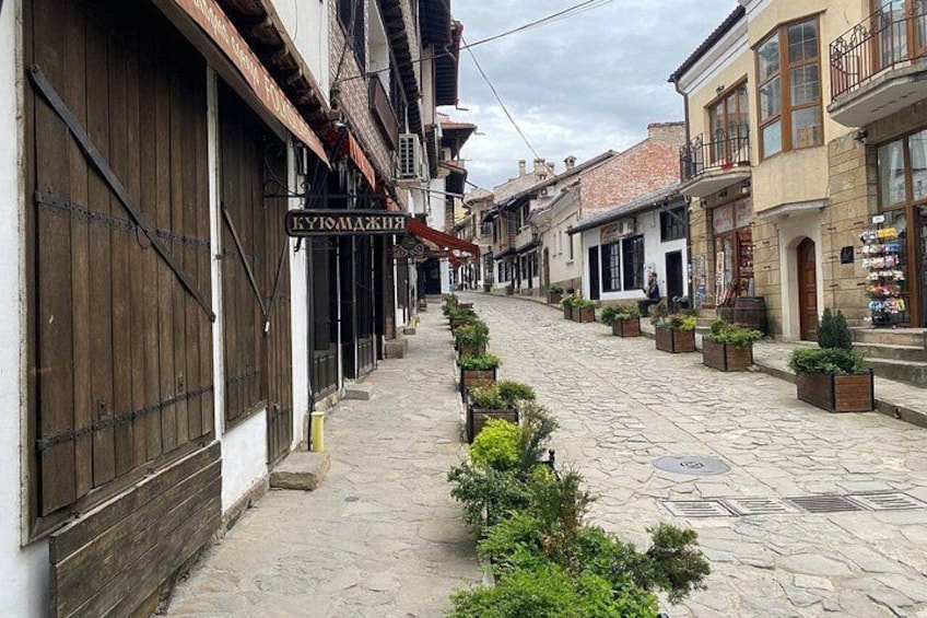 A cobblestone street lined with traditional buildings, a sign reading 'KUKA LUKA,' and potted plants along the sidewalk. 2
