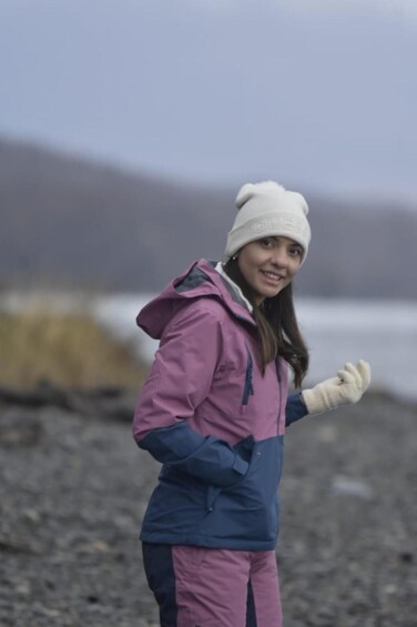 A person wearing a pink and blue jacket and a white beanie, standing on a rocky shore with a body of water and hills in the background. 3