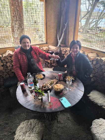 Two people toasting at a wooden table with food and drinks, surrounded by a rustic setting with logs and a wooden bench. 2