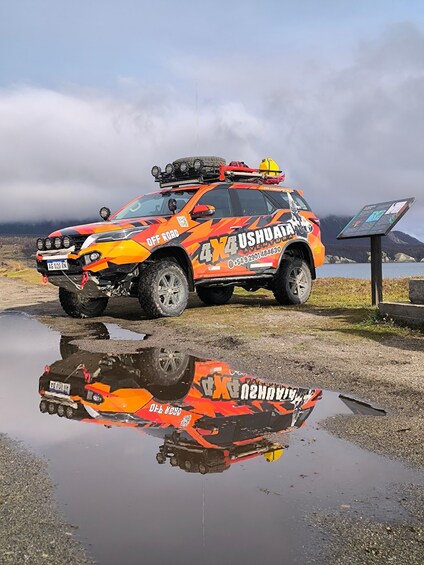 A 4x4 off-road vehicle with 'USHUATAN' branding parked by a reflective puddle, with a mountainous backdrop and a signboard. 1