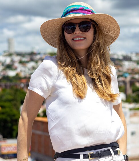 A person wearing a white shirt and a wide-brimmed hat, standing outdoors with a cityscape in the background. 2