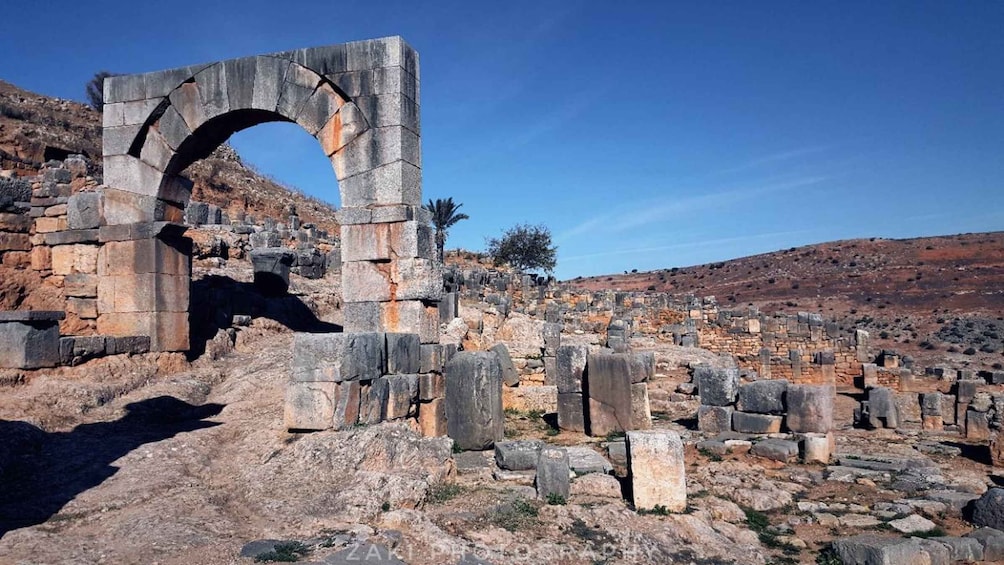 Ancient stone ruins with a large archway, scattered stone blocks, and a hilly landscape in the background. 1