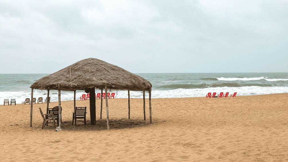 A beach with a thatched hut, wooden chairs, and red plastic chairs. 3