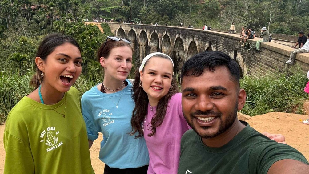 A group of people standing in front of a stone bridge with arches, surrounded by greenery. 2