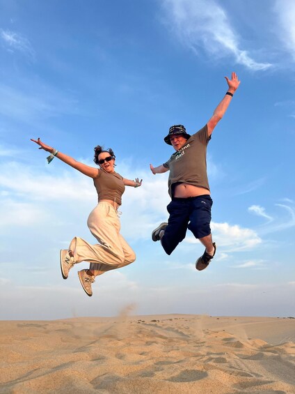 Two people jumping in the air with a clear blue sky and sand dunes in the background. 2