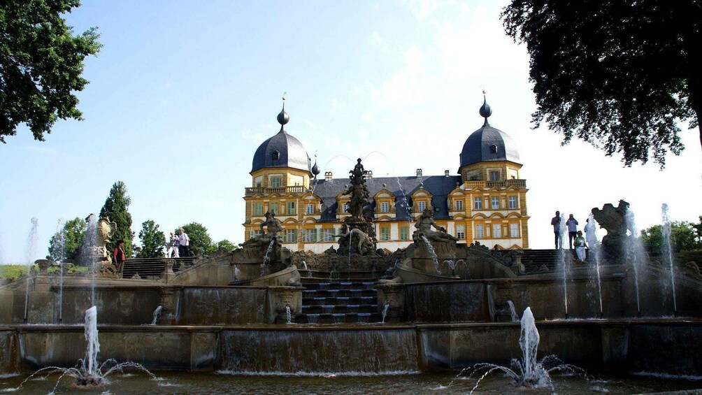A grand building with two domes, a fountain with statues, and people around. 1