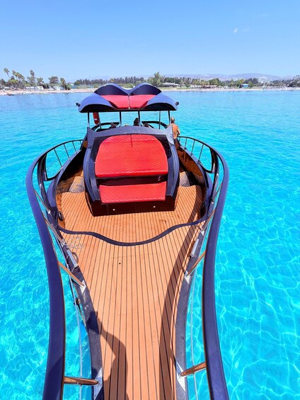 A boat with a red cushioned area and a wooden deck, floating on clear blue water with a beach and palm trees in the background. 4