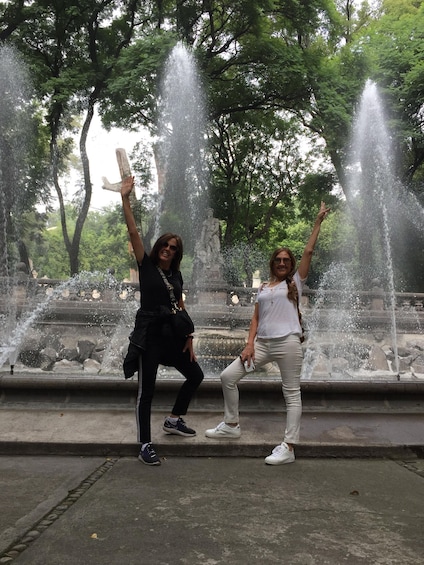 Two people posing in front of a water fountain with multiple streams, surrounded by trees and a stone structure. 2
