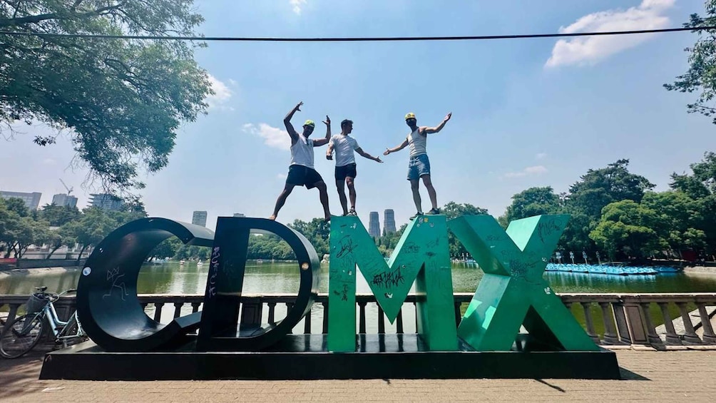 Three people standing on a structure with large letters spelling 'CDMX' in a park with a lake and city buildings in the background. 1