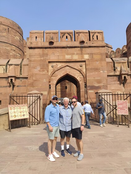 A historical fort with a large arched entrance, a signboard with symbols, and a group of people standing in front. 2