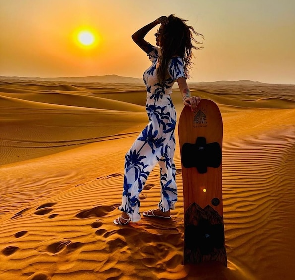 A person standing on a sand dune with a snowboard, with the sun setting in the background. 5