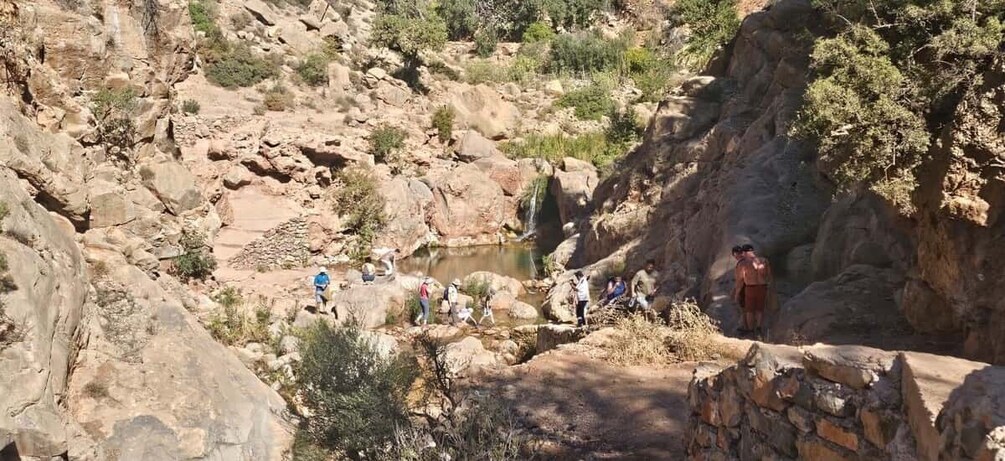 A rocky landscape with a small waterfall and a group of people sitting and standing around a pool. 2
