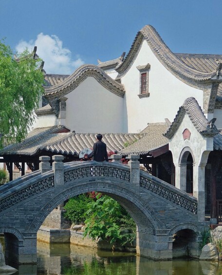 A traditional Chinese bridge over a canal, with a person sitting on the bridge and a building with ornate roof tiles in the background. 2