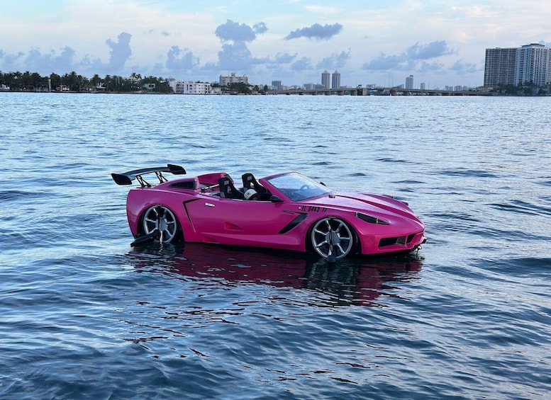 Un'auto sportiva rosa parzialmente immersa in acqua calma, con sullo sfondo la skyline di una città. 2