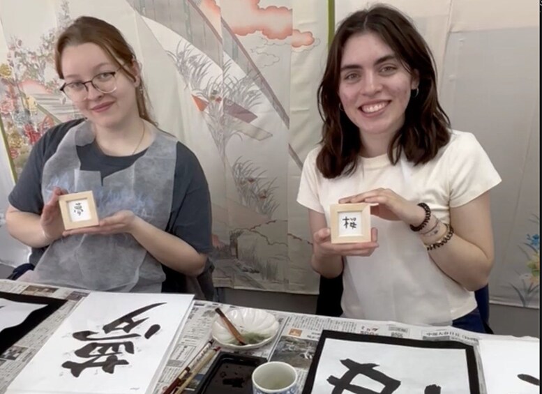 Two individuals holding framed calligraphy pieces at a table with calligraphy and newspapers. 2